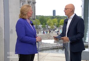 Bärbel Bas beim Sommerinterview im Außenbereich des ARD-Hauptstadtstudios. Im Hintergrund protestiert der Verein Nuklearia e.V. für „klimafreundliche und preiswerte Kernenergie zur Erhaltung von Industriearbeitsplätzen“