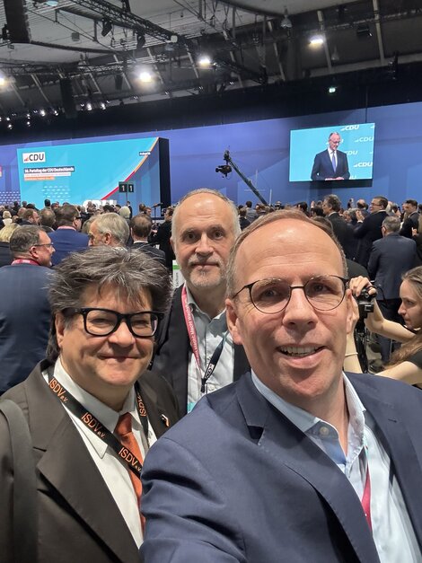 Selfie mit Kanzler: Marcus Pohl (links, isdv und BAGSV), Andreas Lutz (Mitte) und Jörn Freynick (rechts, beide VGSD). Auf der Leinwand: Friedrich Merz.